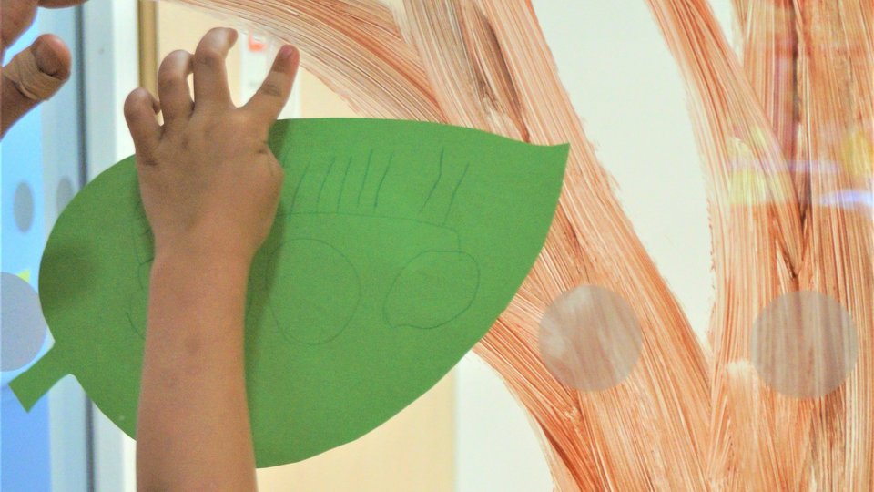 Eine Kinderhand klebt ein grünes Blatt an die Wand. 