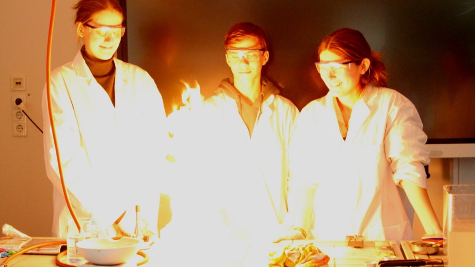 Drei Jugendliche mit weißen Kitteln stehen hinter einer Flamme