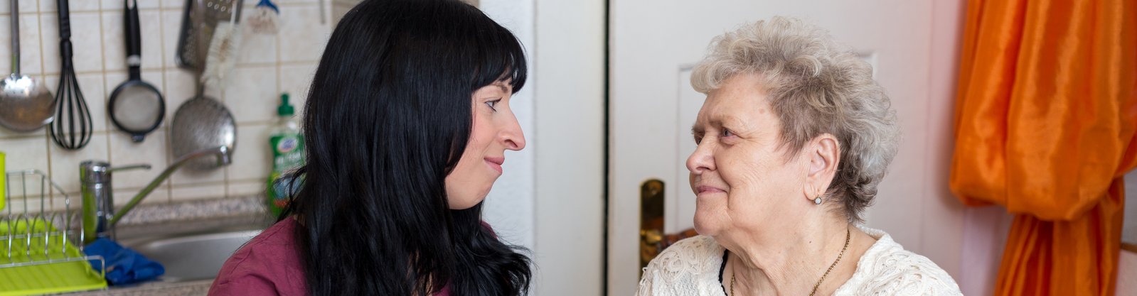 Pflegerin des ambulanten Pflegedienstes bei Seniorin zuhause. Beide schauen sich an und halten sich an den Händen
