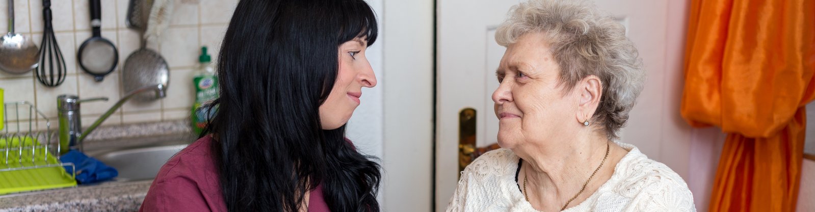 Pflegerin des ambulanten Pflegedienstes bei Seniorin zuhause. Beide schauen sich an und halten sich an den Händen