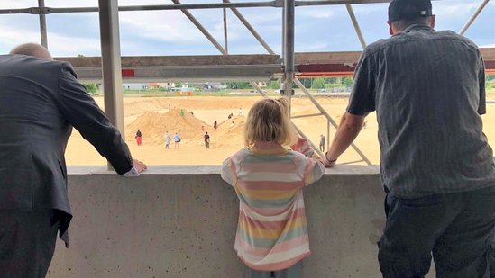 zwei Erwachsende und ein Kind blicken aus dem Gebäude auf die Fläche, auf der Kinder im Sand spielen