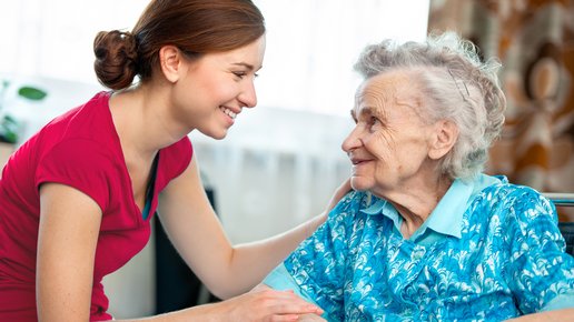 Pflegefachfrau hockt sich zugewandt zu Seniorin. Beide schauen sich freundlich an.