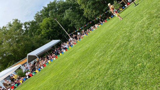 Auf einer Wiese sitzen viele Menschen und hören den Festreden zu