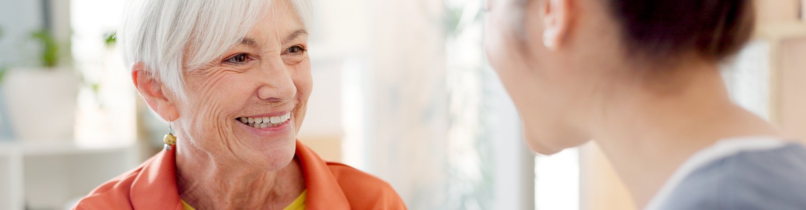 Seniorin lächelt Pflegekraft an. Beide geben sich vertraut die Hand.