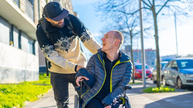 Zwei Männer beim Spaziergang, ein Betreuer, der den anderen körperlich beeinträchtigt und im Rollstuhl sitzend schiebt